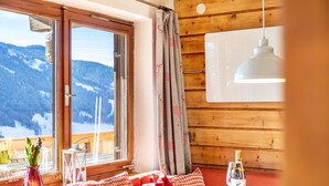 A wooden cabin with a dining area, a window with a view of snow-covered mountains, and a table with a bottle and glass.