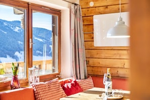 A wooden cabin with a dining area, a window with a view of snow-covered mountains, and a table with a bottle and glass.