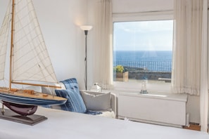 Ein Zimmer mit einem Bett, einem Modell eines Segelbootes, einer Lampe und Blick auf das Meer durch ein Fenster.