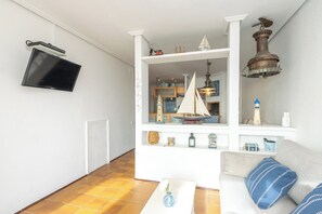 Ein Wohnzimmer mit an der Wand montiertem Fernseher, einem Sofa mit blauen Kissen, einem kleinen Tisch mit einer Vase und einem Regal mit verschiedenen Dekorationsobjekten.