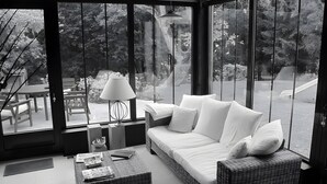Une pièce moderne avec un canapé blanc, un pouf en osier et une table avec une lampe. De grandes fenêtres offrent une vue sur un jardin et des arbres.