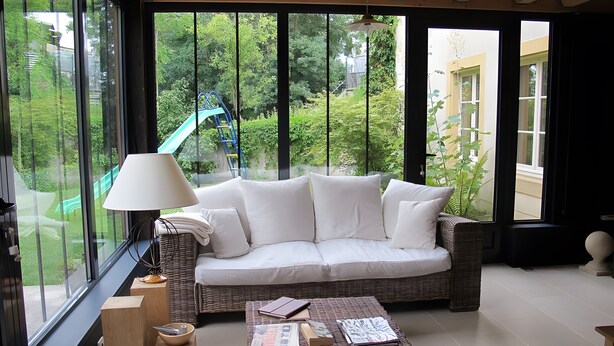 Une pièce avec un canapé blanc, une table basse en osier, une lampe et une vue sur la verdure grâce à de grandes fenêtres.