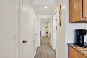 A narrow hallway with white walls and wooden flooring, leading to a kitchen area with a coffee maker on the counter. - Enjoy your stay at this fully equipped first-floor beachfront condo, with a pool (Tybee Island)