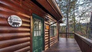 En stuga byggd av trä med en grön dörr, en välkomstskylt och en veranda med utsikt över ett skogsområde.