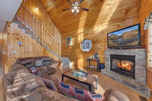 A cozy cabin interior with a fireplace, a television mounted on the wall, a staircase, and a large window. - 2 Kings and a Sofa Sleeper Hot Tub Near Dollywood (Sevierville)