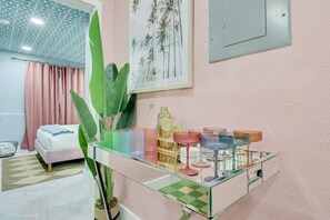 A hotel room with a bed, a glass side table with colorful glassware, a large green plant, and a wall-mounted light switch. - Hot Tub + Makeup Bar + Karaoke + Design District (Miami)