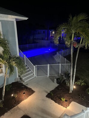 A nighttime view of a residential backyard with a swimming pool, palm trees, and a white fence. - The Finn at Coconut Cove (Fort Myers Beach)