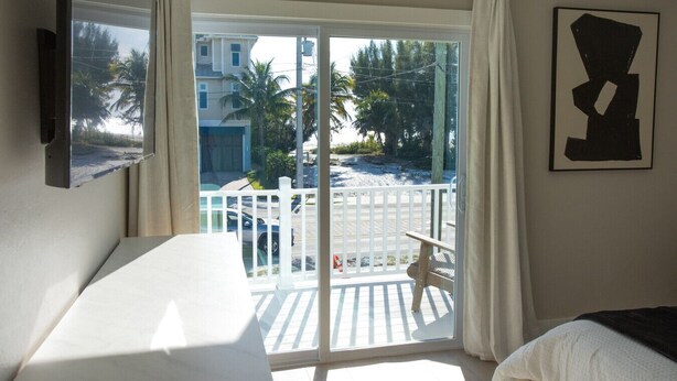 A hotel room with a bed, a TV mounted on the wall, a balcony with a white railing, and a view of palm trees and buildings outside. - The Finn at Coconut Cove (Fort Myers Beach)