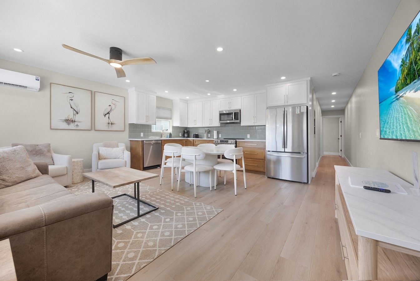 Cocina moderna con armarios blancos, comedor con mesa y sillas blancas, y sala de estar con sofá color beige.