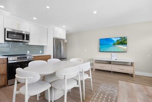 Une cuisine moderne avec des meubles blancs, un réfrigérateur en acier inoxydable et un four à micro-ondes. On y trouve une table à manger avec des chaises blanches et un téléviseur fixé au mur.