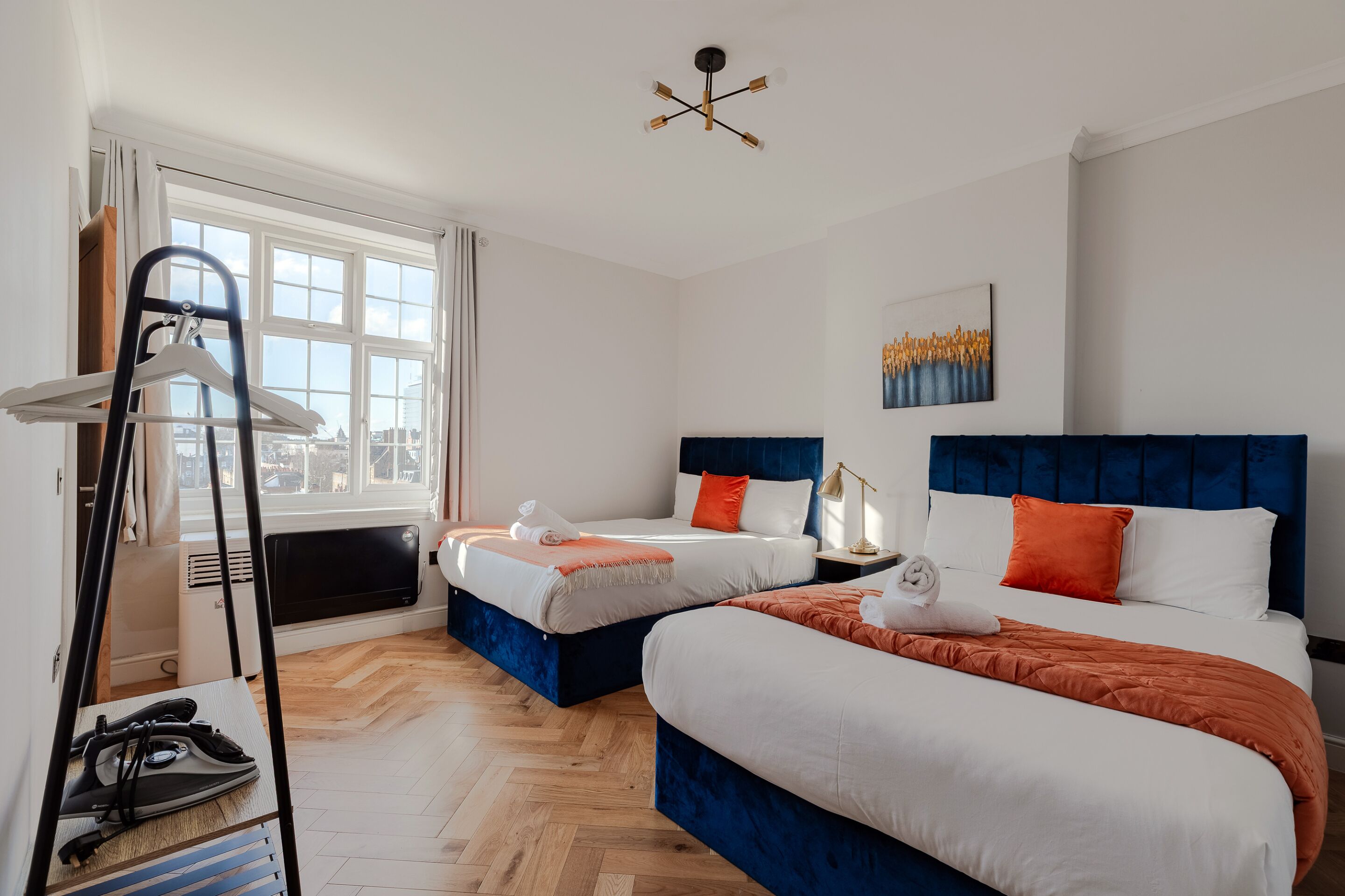 A hotel room with two beds, a large window, a radiator, and a ceiling light fixture.