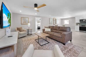 A modern living room with a sofa, armchairs, a coffee table, and a TV. There is a ceiling fan, a kitchen in the background, and a patterned rug. - The June at Coconut Cove (Fort Myers Beach)