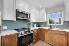 A modern kitchen with white cabinets, a stainless steel oven, a microwave, and a window with a view. - The Dex at Coconut Cove (Fort Myers Beach)