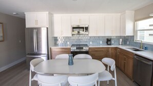 A modern kitchen with white cabinets, stainless steel appliances, a round dining table with chairs, and a tiled backsplash. - The Wren at Coconut Cove (Fort Myers Beach)
