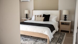 A bedroom with a bed, two bedside tables, a wall-mounted air conditioner, and a window with blinds. - The Wren at Coconut Cove (Fort Myers Beach)