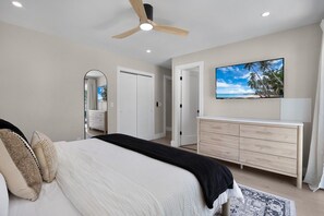 A bedroom with a bed, a dresser, a ceiling fan, a TV, and a door leading to another room. - The Wren at Coconut Cove (Fort Myers Beach)