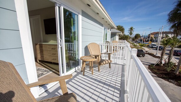 A balcony with chairs and a wooden cabinet, a white railing, and a view of the street with palm trees and parked cars. - The Wren at Coconut Cove (Fort Myers Beach)