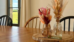 Ein Holztisch mit zwei Glasvasen, gefüllt mit getrockneten Blumen, ein gerahmtes Bild an der Wand und Blick auf ein grünes Feld durch ein Fenster.