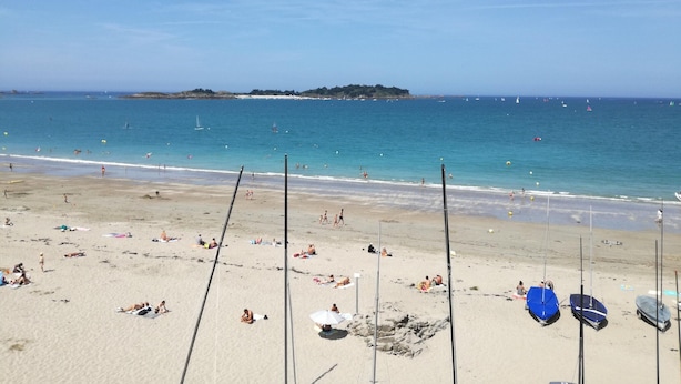 Ein Strand mit badenden und sonnenbächelnden Menschen, Booten, die in der Nähe der Küste ankerten, und einer entfernten Insel.