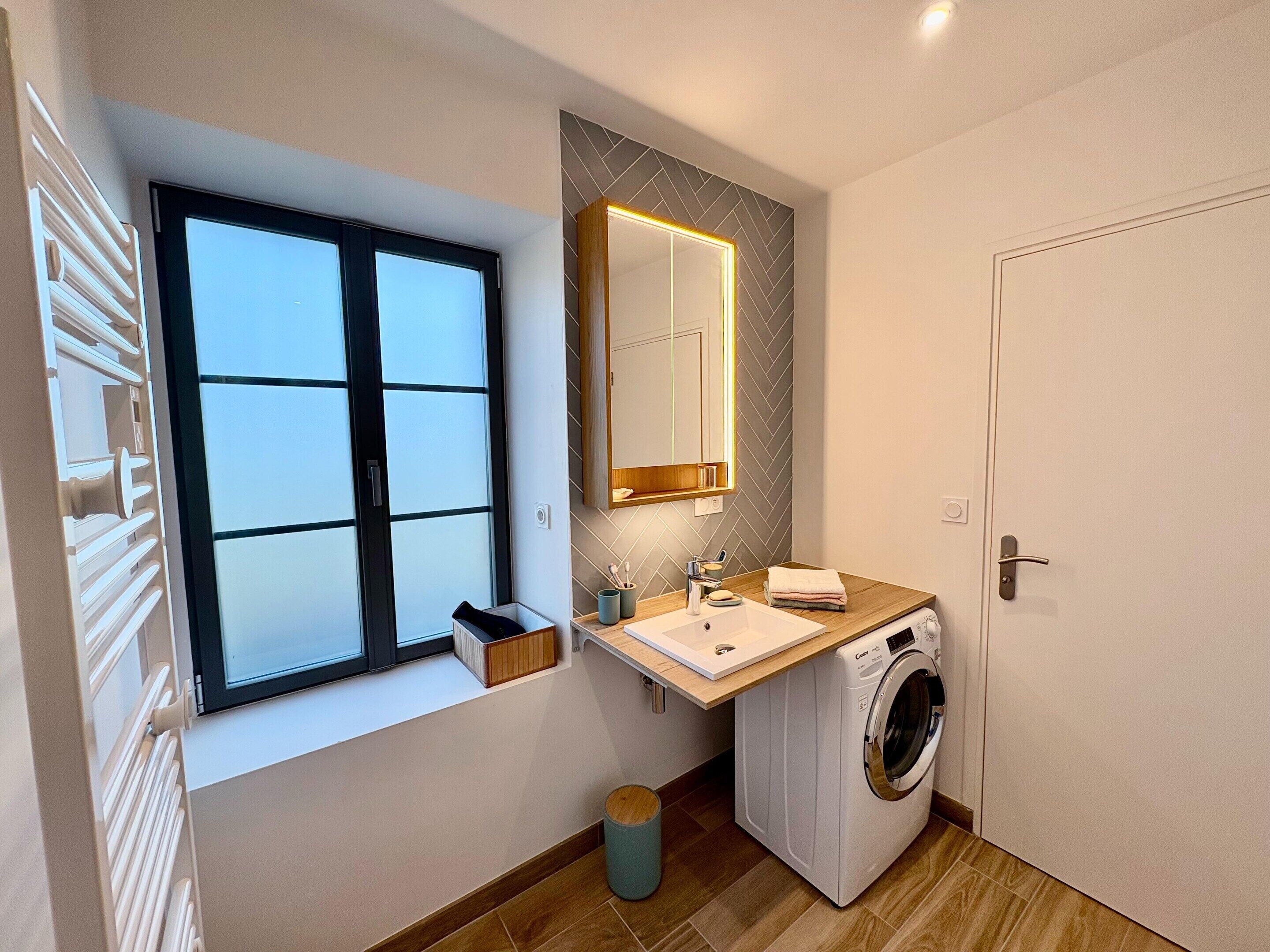 A compact bathroom with a window, a built-in washing machine, and a mirror with a wooden frame.