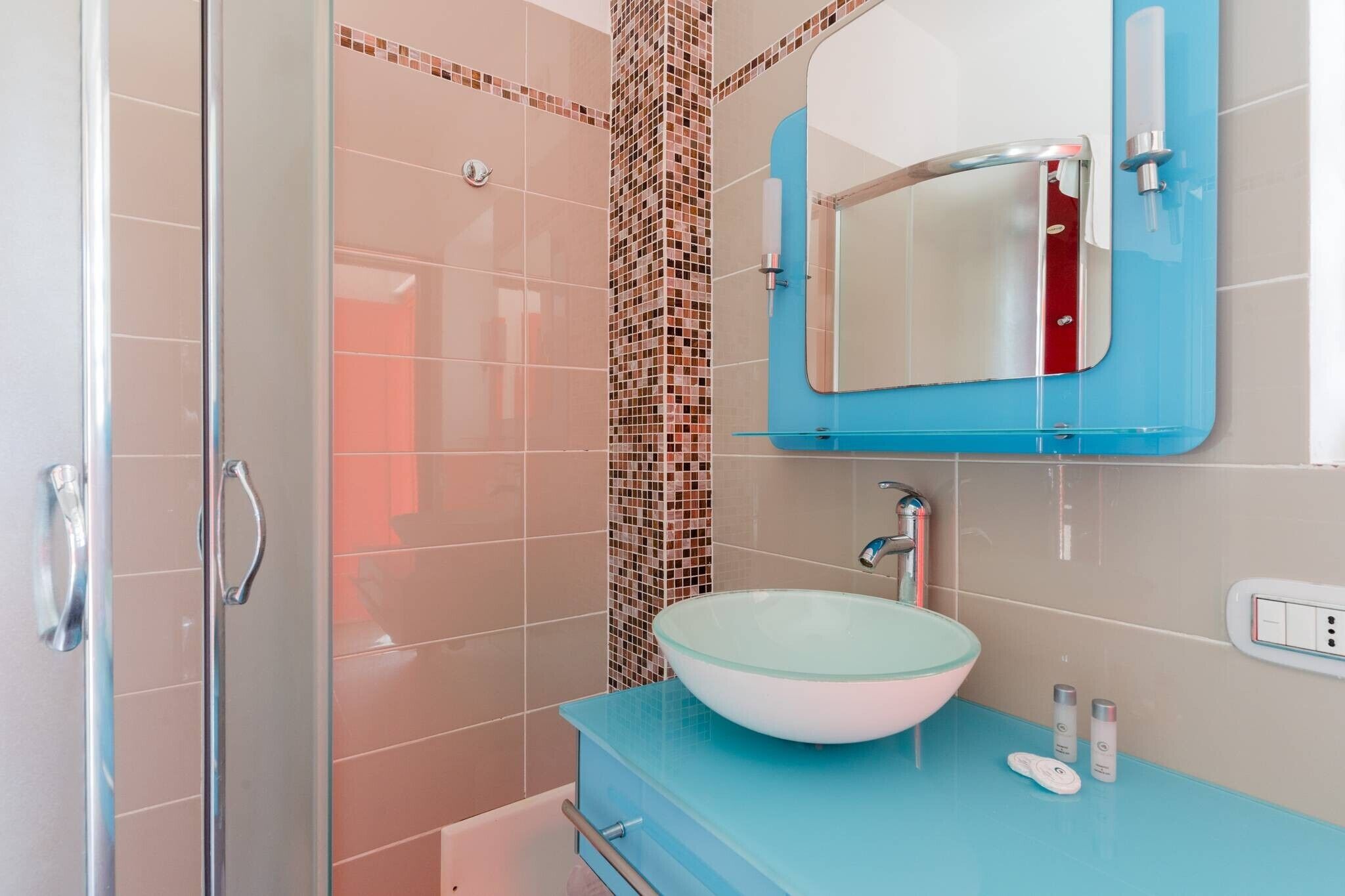A bathroom with a glass sink, a blue vanity, a shower with a glass door, and a tiled wall with a mosaic pattern.