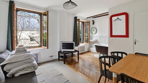 Una sala de estar moderna con un televisor sobre un mueble bajo, una mesa de comedor con sillas, un sofá con una pila de mantas y ventanas amplias con cortinas.