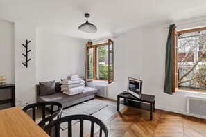 A modern living room with a sofa, a dining table, chairs, a TV, and large windows with curtains. - Premium 1BR flat with direct access to the RER B (Gentilly)