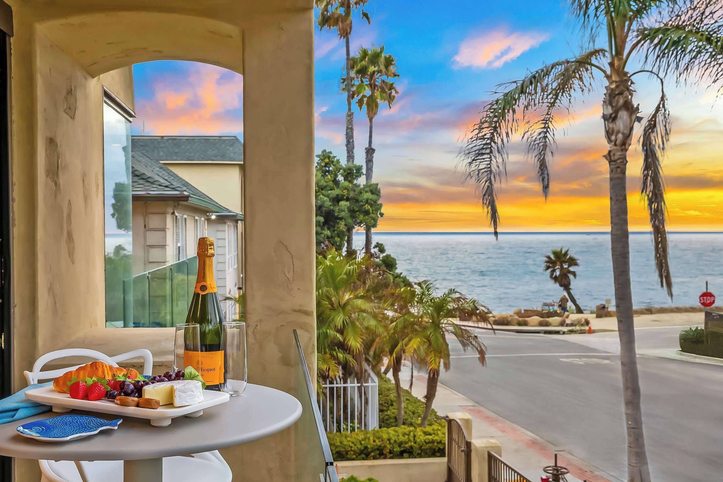 A balcony with a table set for a meal, overlooking a sunset view with palm trees and a beach.
