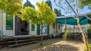 A mobile home with a covered outdoor area, gravel ground, and a small table with chairs. - “3-Bedroom Mobile Home in St Aygulf” with Shared Pool, Wi-Fi, and Air Conditioning (Fréjus)