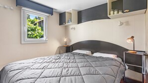A bedroom with a large bed, a window with a view of greenery, a bedside table with a lamp, and a built-in shelf above the bed. - “3-Bedroom Mobile Home in St Aygulf” with Shared Pool, Wi-Fi, and Air Conditioning (Fréjus)