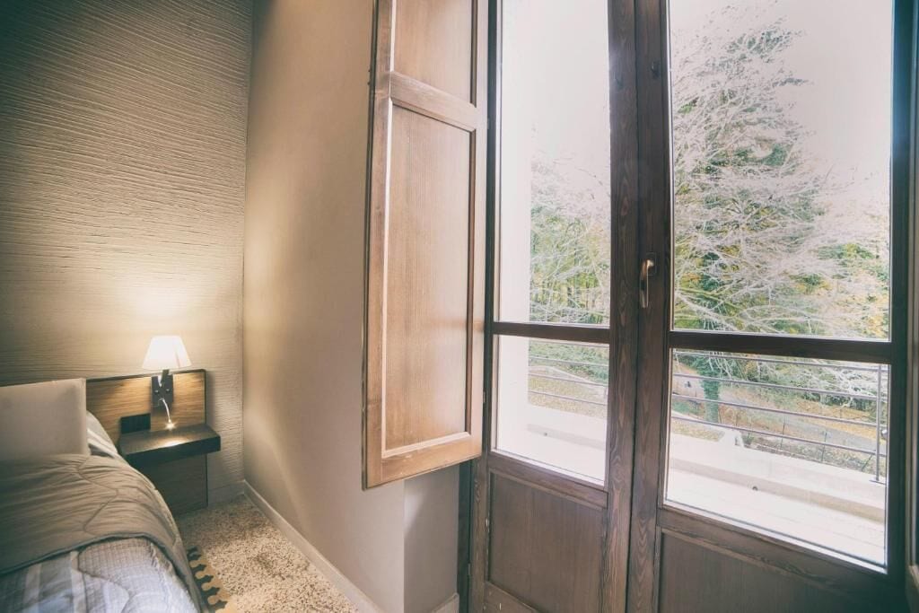Ein Schlafzimmer mit einem Bett, einem Nachttisch mit Lampe, einem Fenster mit Blick auf Bäume und einer Tür.