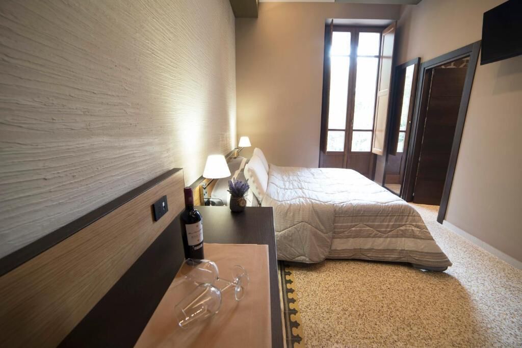 A hotel room with a bed, a bedside table with a wine bottle and glasses, a lamp, and a view of the outside through a window.