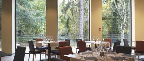 A restaurant with tables set for dining, large windows with a forest view, and a modern interior design.