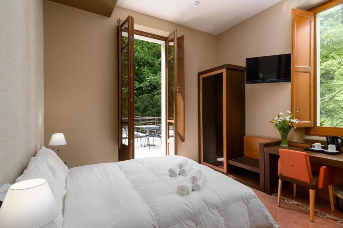 A hotel room with a bed, a desk, a chair, a TV, and a window with a view of greenery.