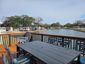 A waterfront deck with tables and chairs, a view of the water, and RVs parked along the shore.
