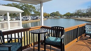 A wooden deck with two chairs and a table, overlooking a calm body of water with RVs parked nearby.
