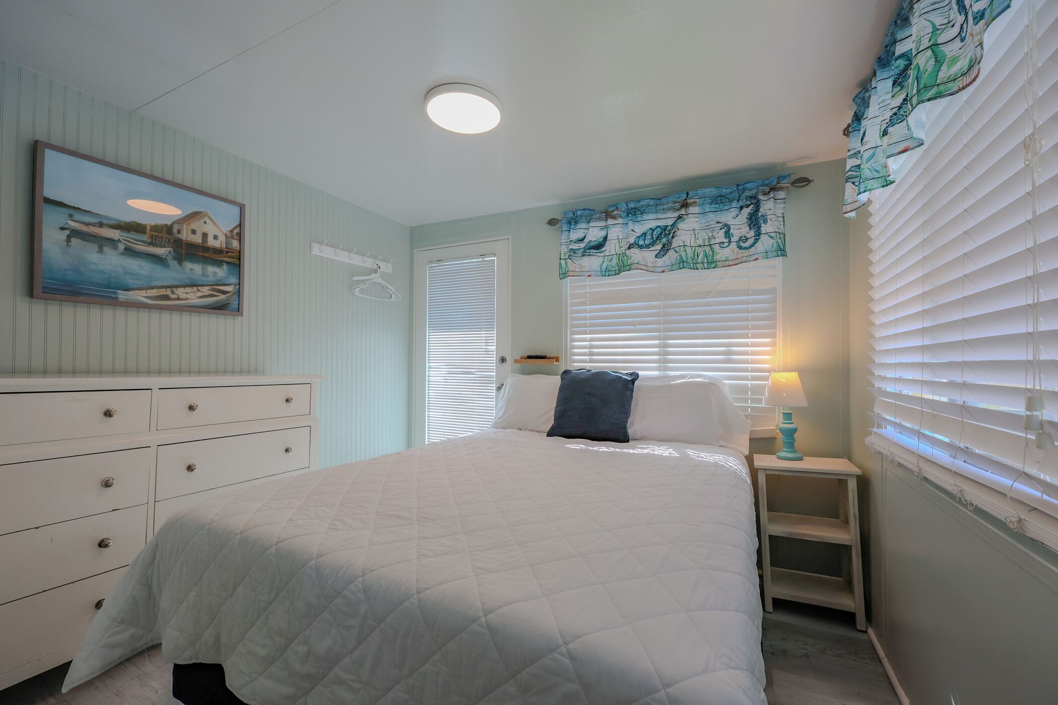 A bedroom with a bed, a dresser, a nightstand, a lamp, a window with blinds, and a painting on the wall.