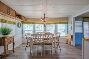 Un espace repas comprenant une table et des chaises en bois, un lustre et une vue sur l’extérieur grâce à de grandes fenêtres.