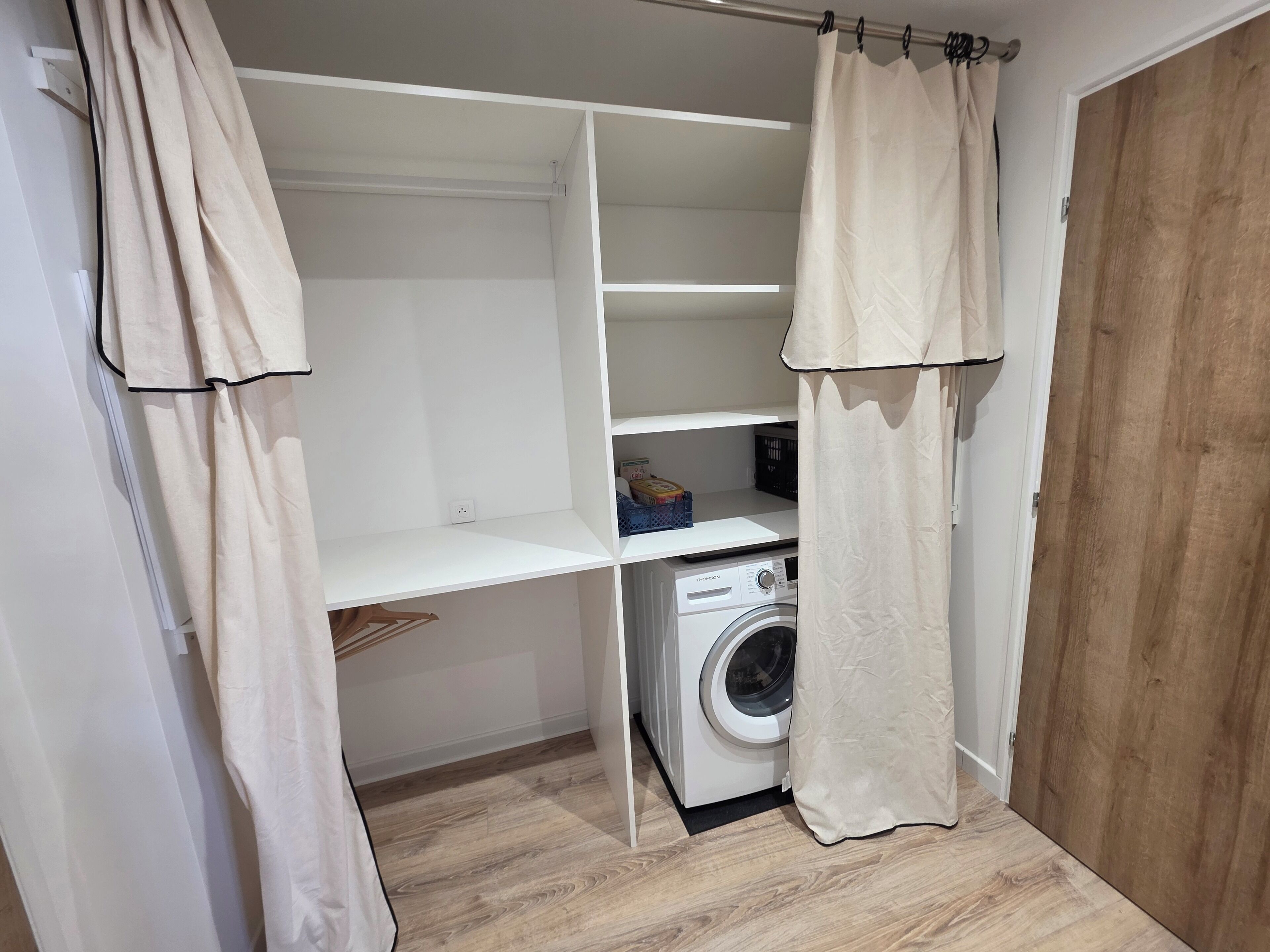 A laundry room with a washing machine, shelves, and a wooden floor.