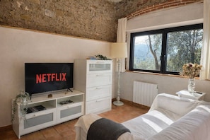 A living room with a television displaying 'NETFLIX,' a white sofa, a white cabinet, a floor lamp, and a window with a view of trees. - Adults Only Rural | 2 P | Pool | WiFi| BBQ| Girona (Madremanya)