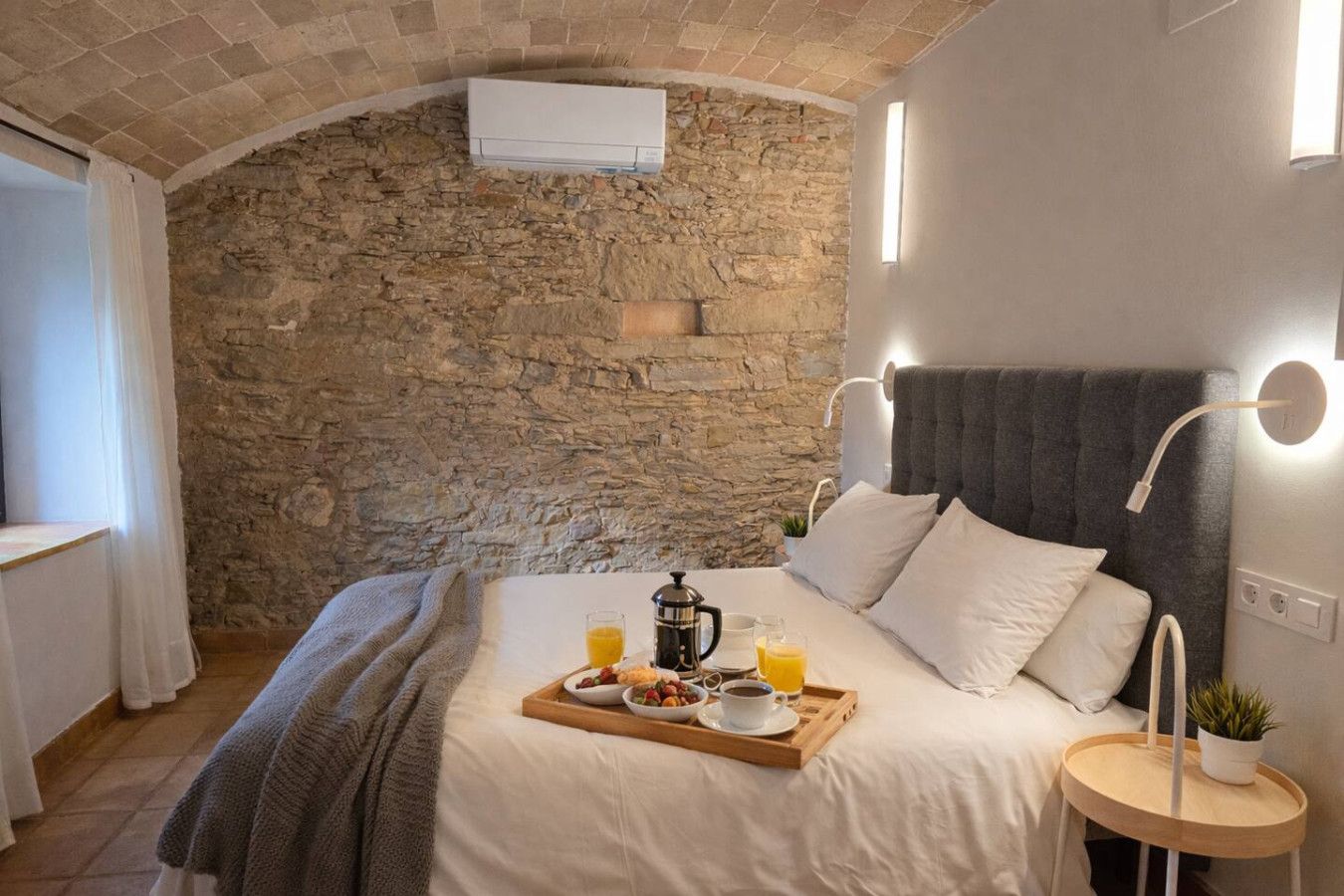 A bedroom with a stone wall, a bed with white bedding, a tray with breakfast, and a bedside table with a plant.