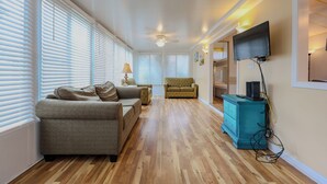 A spacious living room with hardwood flooring, a ceiling fan, and a television mounted on the wall.