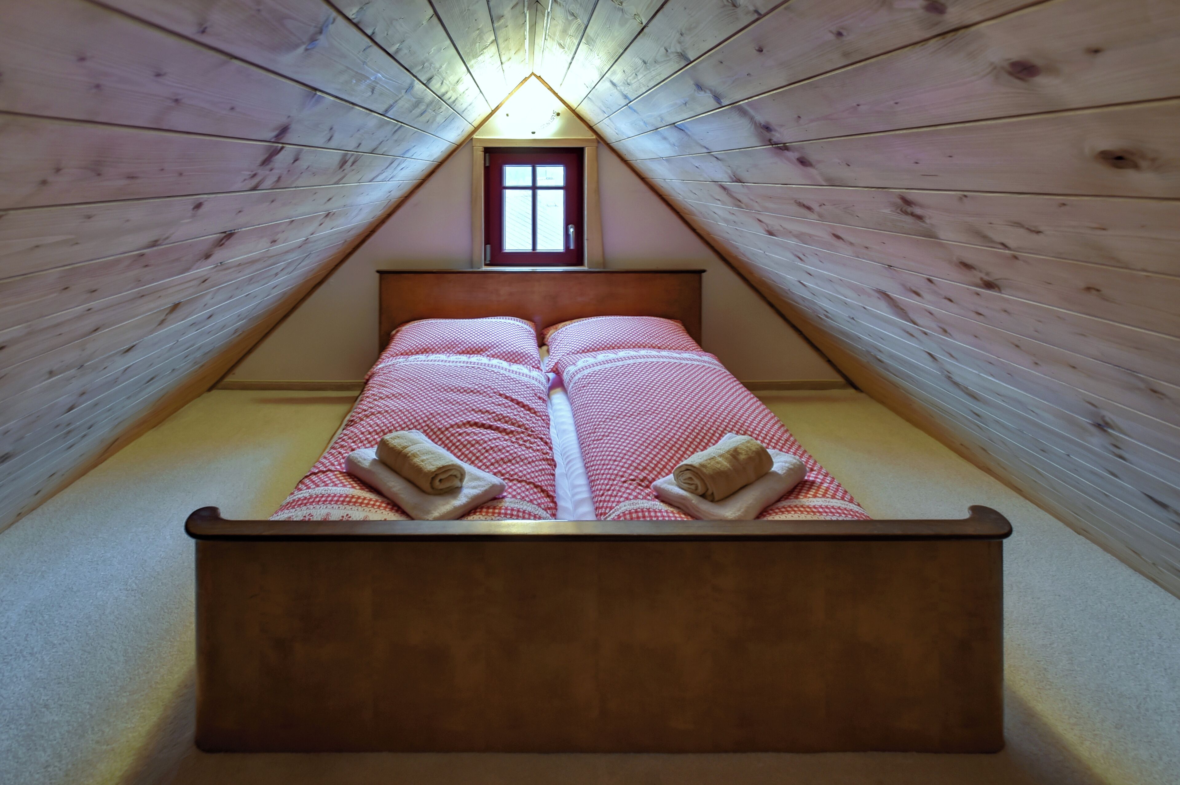 A bedroom with a wooden sloped ceiling, a small window, and a bed with red and white checkered bedding and rolled towels.