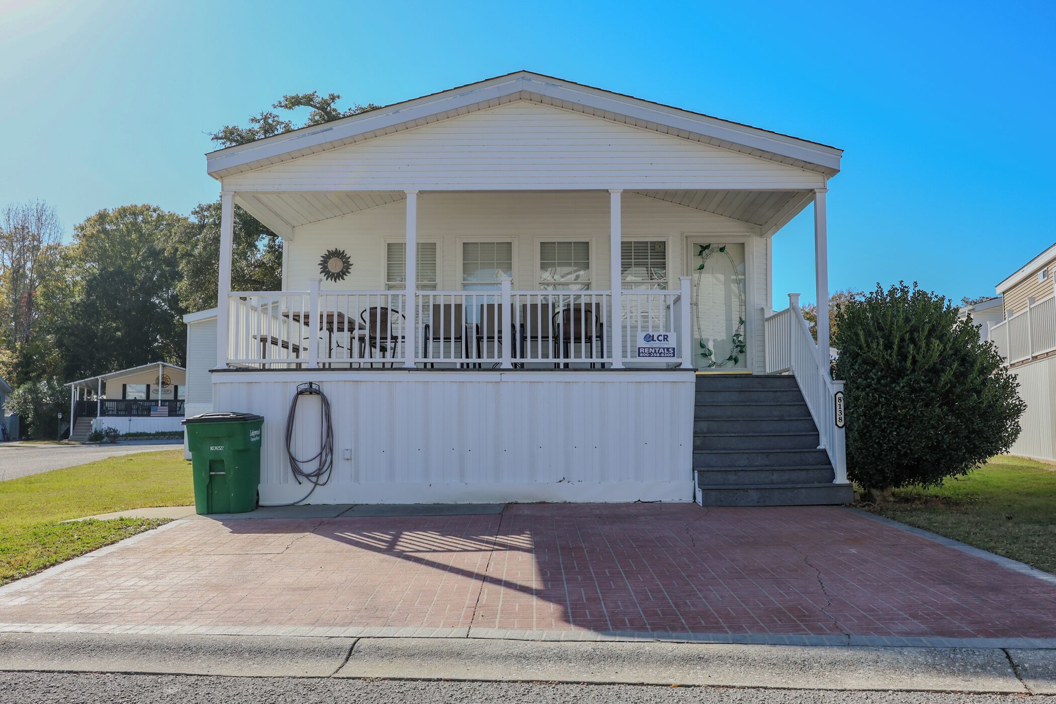 A white single-story house with a porch, a small set of stairs leading to the entrance, and a green trash bin.
