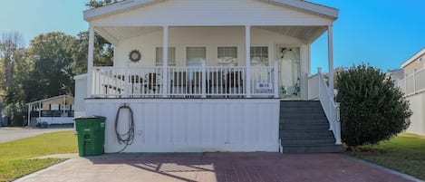 A white single-story house with a porch, a small set of stairs leading to the entrance, and a green trash bin.
