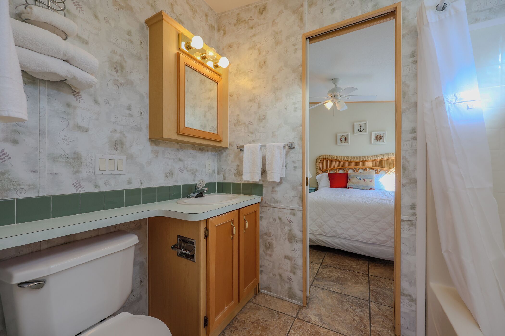 Une salle de bain équipée de toilettes, d’un lavabo et d’un miroir avec éclairage. On peut apercevoir un lit à travers une porte ouverte.