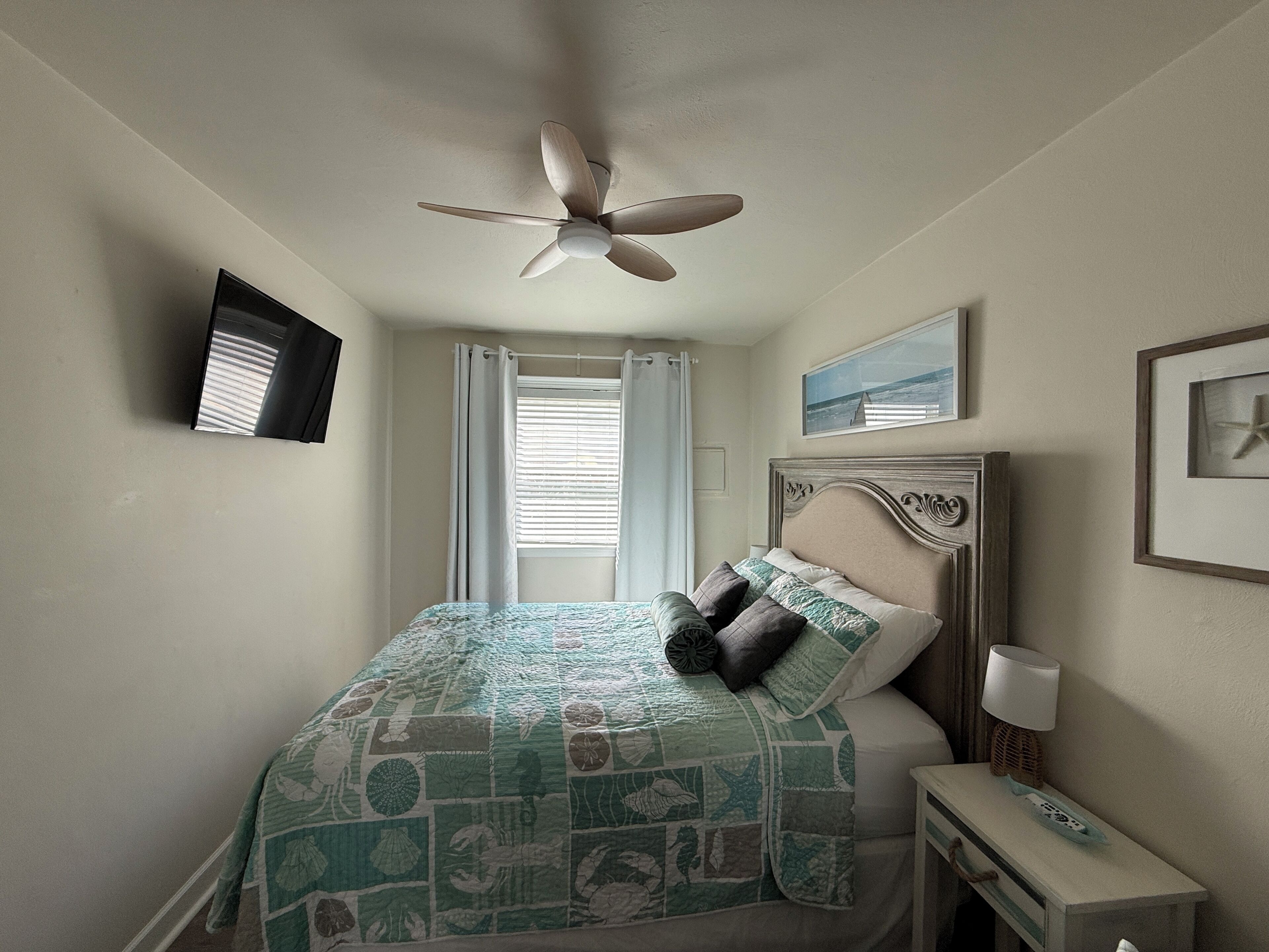 A bedroom with a bed, a ceiling fan, a window with curtains, a bedside table with a lamp, and a framed picture on the wall.