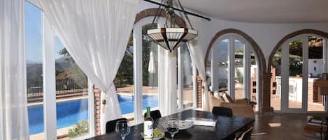 A dining area with a view of a swimming pool, large windows, and a hanging light fixture.