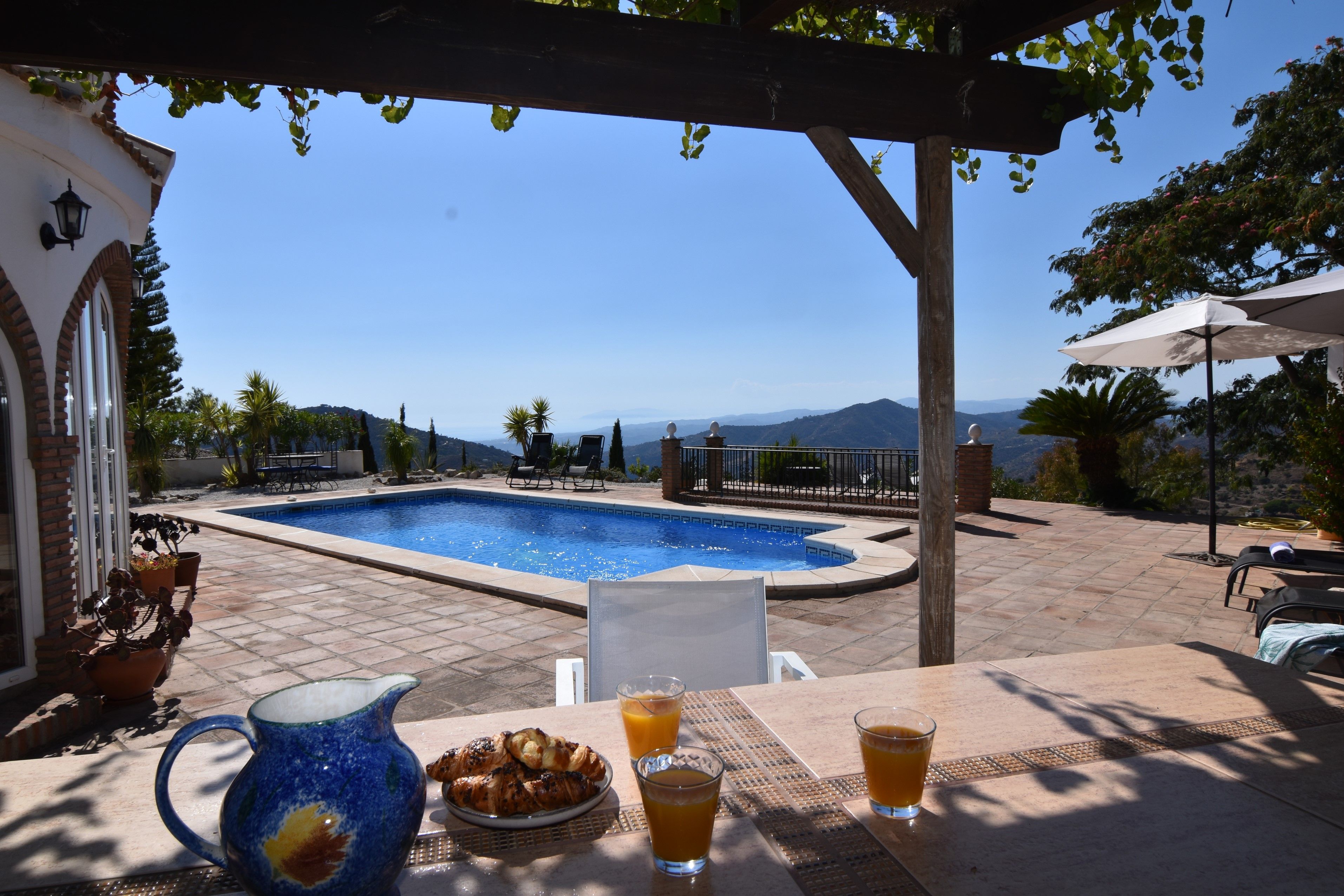 A poolside area with a view of mountains, a table with breakfast, and a pergola with hanging plants.