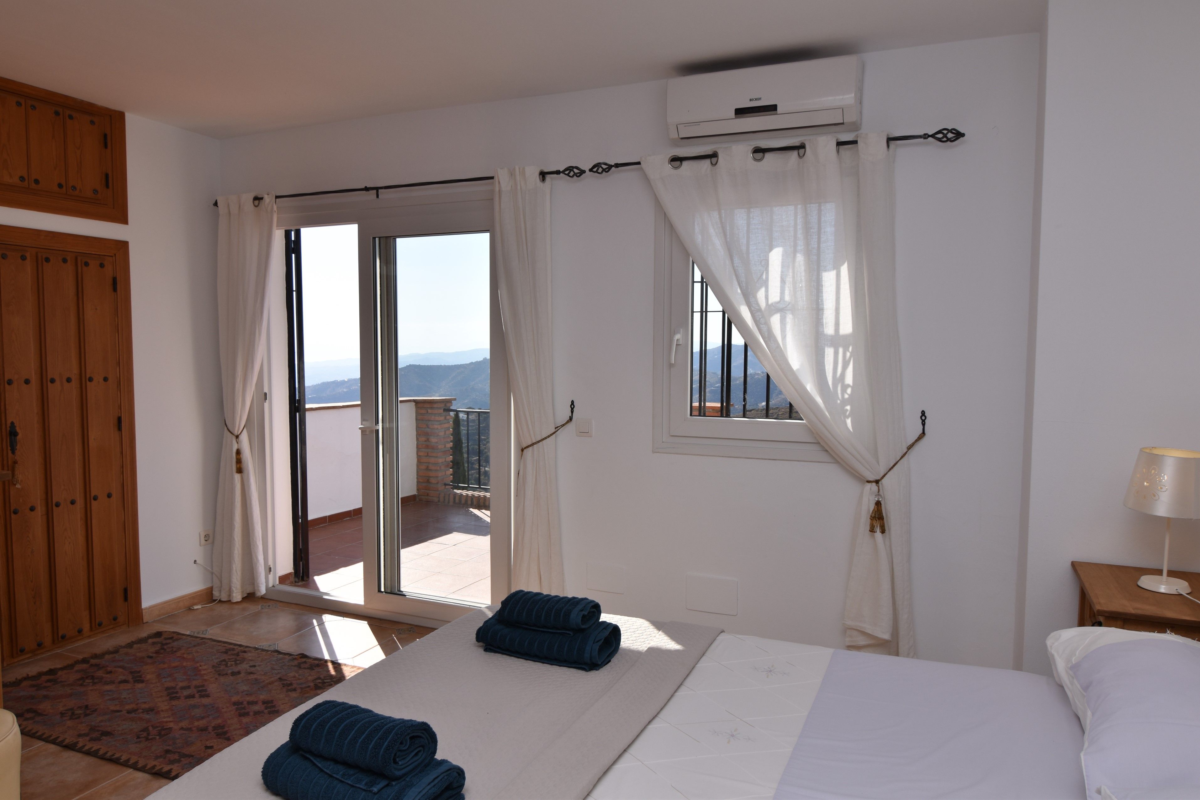 A bedroom with a bed, a window with curtains, a wooden wardrobe, and a view of the outdoors.
